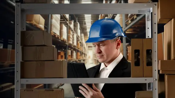 Business warehouse and logistics creative advertisement concept. Portrait of storekeeper working in storage. Man manager in formal outfit in blue helmet stands checks packages on rack with goods.