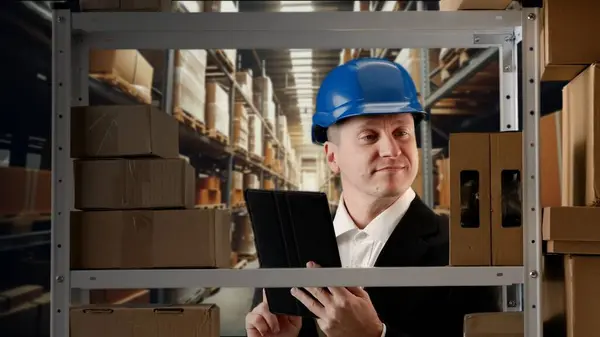 Business warehouse and logistics creative advertisement concept. Portrait of storekeeper working in storage. Man manager in formal outfit in blue helmet stands checks packages on rack with goods.