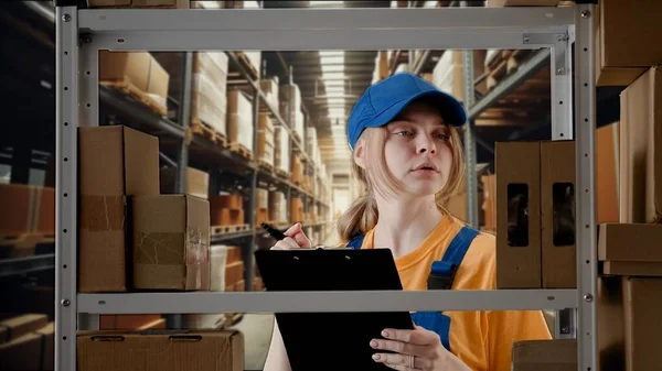 Business warehouse and logistics creative advertisement concept. Portrait of female model working in storage. Girl storekeeper in uniform standing near rack with boxes, checks goods writing in blank.