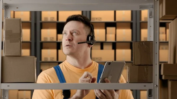 Business warehouse and logistics creative advertisement concept. Portrait of male working in storage. Man storekeeper standing near rack in headset talking and checking goods availability on tablet.