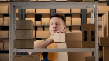 İş deposu ve lojistik yaratıcı reklam konsepti. Depoda çalışan erkek portresi. Erkek mağazacı rafın yanında duruyor. Elinde büyük bir deste olan kutuları alıp paketleri bırakıyor..