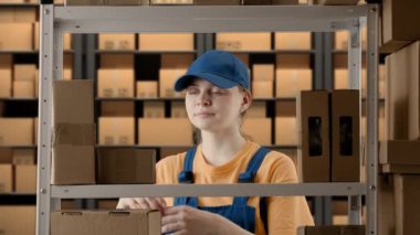 İş deposu ve lojistik yaratıcı reklam konsepti. Depoda çalışan kadın modelin portresi. Üniformalı kız mağaza görevlisi kutularla rafın yanında duruyor, paket gösteriyor ki başparmak kaldırıyor..