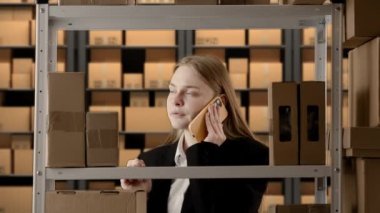 İş deposu ve lojistik yaratıcı reklam konsepti. Depoda çalışan bir kadının portresi. Kız mağaza müdürü resmi kıyafet içinde kutuların yanında, akıllı telefondan konuşuyor..