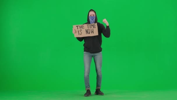スカーフで覆われた顔をした男性のデモンストレーションは 時が今であるというサインを保持します 男は緑色のスクリーンでスローガンを叫ぶ クロマキー — ストック動画