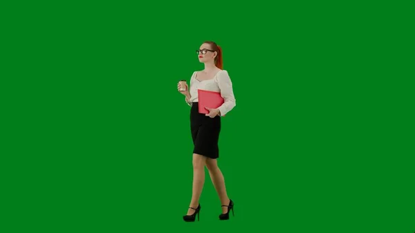 Modern business woman creative advertisement concept. Portrait of attractive office girl on chroma key green screen. Woman in skirt and glasses walking holding red folder and coffee cup. Half turn.