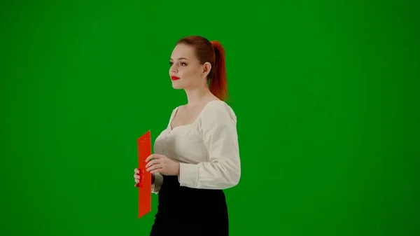 Modern business woman creative advertisement concept. Portrait of attractive office girl on chroma key green screen. Woman in skirt and blouse walking holding red folder. Half turn.
