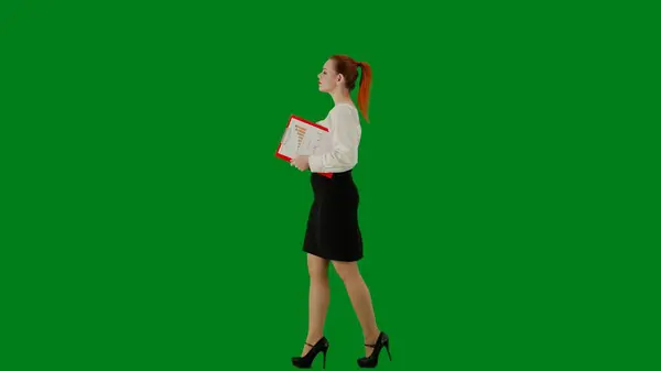Modern business woman creative advertisement concept. Portrait of attractive office girl on chroma key green screen. Woman in skirt and blouse walking holding red folder. Side view.