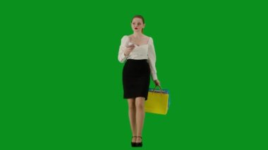 Modern iş kadını yaratıcı reklam konsepti. Krom anahtar yeşil ekranda çekici ofis kızı portresi. Etekli bir kadın elinde hediye çantalarıyla yürüyor ve akıllı telefondan mesaj atıyor, gülümsüyor..