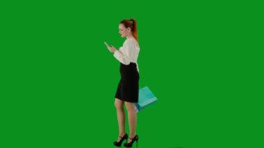 Modern iş kadını yaratıcı reklam konsepti. Krom anahtar yeşil ekranda çekici ofis kızı portresi. Etekli bir kadın elinde hediye çantalarıyla yürüyüp akıllı telefondan mesaj atıyor. Yan görünüm.