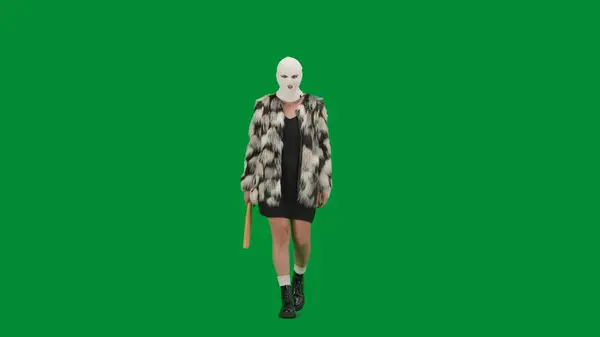 Woman in white balaclava, fur coat and evening dress walking with bat in hand. Woman freak on green background in studio. Fashion trend concept, feminist trend in fashion