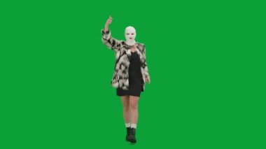 Beyaz kar maskeli kadın, kürk manto ve gece elbisesi giymiş elleri havada neşeyle yürüyor ve dans ediyor. Stüdyoda yeşil arka plandaki kadın ucubesi. Moda eğilimi, feminist moda eğilimi