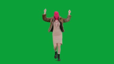 Pembe kar maskeli kadın, kaplan kürkü manto ve gece elbisesi giymiş elleri havada neşeyle yürüyor ve dans ediyor. Stüdyoda yeşil arka plandaki kadın ucubesi. Moda eğilimi, feminist moda eğilimi