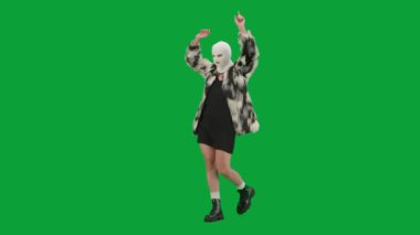 Beyaz kar maskeli kadın, kürk manto ve gece elbisesi giymiş elleri havada neşeyle yürüyor ve dans ediyor. Stüdyoda yeşil arka plandaki kadın ucubesi. Moda eğilimi konsepti Yarım dönüş.