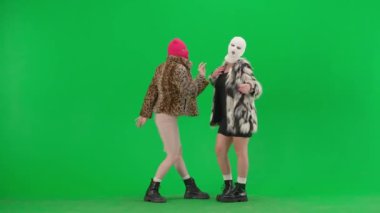 Beyaz ve pembe kar maskeli iki kadın kamera önünde model gibi poz veriyor. Stüdyoda yeşil arka planda kürklü kadınlar. Moda eğilimi, feminist moda eğilimi