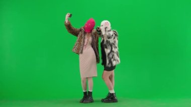 Beyaz ve pembe kar maskeli iki kadın akıllı telefon kullanarak selfie çekiyor. Yeşil arka plan stüdyosunda kürklü kadınlar. Moda eğilimi, feminist moda eğilimi