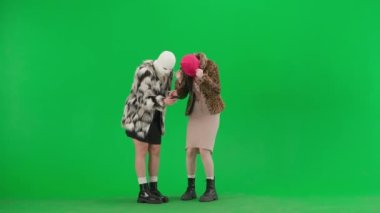 Beyaz ve pembe kar maskeli iki kadın akıllı telefona umutla bakıyor ve iyi haber ya da zafere seviniyor. Yeşil stüdyo arka planında kürk giyen ucube kadınlar. Moda eğilimi konsepti.