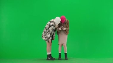 Beyaz ve pembe kar maskeli iki kadın akıllı telefondan oyun oynuyorlar ve zaferden zevk alıyorlar. Yeşil arka plan stüdyosunda kürklü kadınlar. Moda eğilimi, feminist moda eğilimi