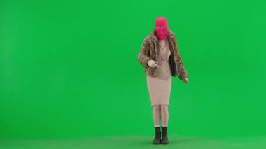 Pembe kar maskeli kadın, kaplan ceketli ve gece elbisesi giyen kadın parmaklarını kenarı işaret ediyor ve baş parmak işareti gösteriyor. Stüdyoda yeşil arka planda bir ucube kadın.