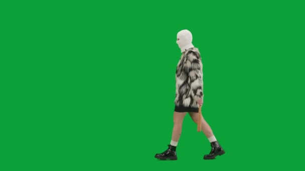 身穿白色巴拉克拉法帽 皮大衣和晚礼服的女人手拿着球棒走着 工作室里的女性对绿色背景非常好奇 时尚潮流的概念 时尚方面的女性主义趋势 — 图库视频影像