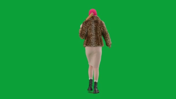身穿粉色巴拉克拉瓦的女人 身穿虎皮大衣 身穿晚礼服 手里拿着球棒走路 工作室里的女性对绿色背景非常好奇 时尚潮流的概念 时尚后视镜中的女性主义趋势 — 图库视频影像