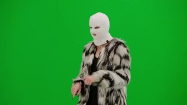 Beyaz kar maskeli kadın, palto ve gece elbisesi giyen dans eden ve işaret parmağını farklı yönlere çeviren kadın. Stüdyoda yeşil arka plandaki ucube kadın. Yarım dönüş..