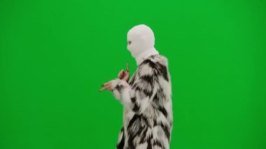 Beyaz kar maskeli kadın, palto ve gece elbisesi giyen dans eden ve işaret parmağını farklı yönlere çeviren kadın. Stüdyo manzaralı yeşil arka planda ucube bir kadın.