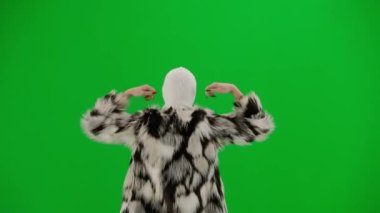 Beyaz kar maskeli, kürklü ve elbiseli bir kadın elleri havada yürüyor ve neşeyle dans ediyor. Stüdyoda yeşil arka plandaki kadın ucubesi. Moda eğilimi kavramı, feminist moda eğilimi