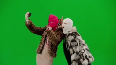 Beyaz ve pembe kar maskeli iki kadın akıllı telefon kullanarak selfie çekiyor. Yeşil arka plan stüdyosunda kürklü kadınlar. Moda eğilimi, feminist moda eğilimi