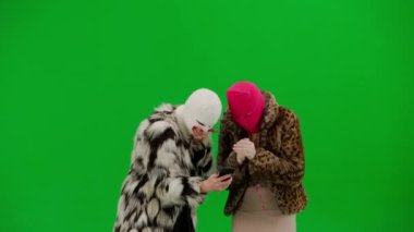 Beyaz ve pembe kar maskeli iki kadın akıllı telefona umutla bakıyor ve bir kayıp ya da kötü haber yüzünden üzülüyorlar. Stüdyonun yeşil arka planında kürk giyen kadınlar.