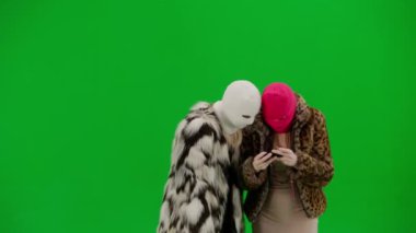 Beyaz ve pembe kar maskeli iki kadın akıllı telefondan oyun oynuyorlar ve zaferden zevk alıyorlar. Yeşil arka plan stüdyosunda kürklü kadınlar. Moda eğilimi, feminist moda eğilimi