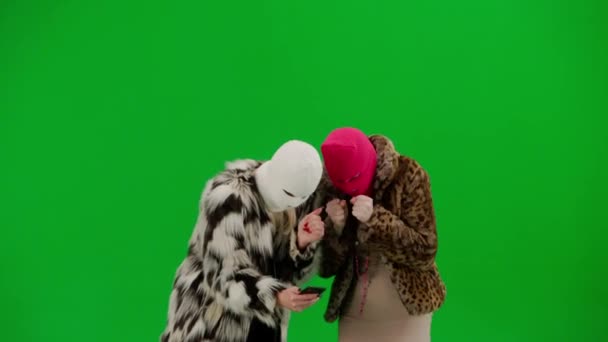 两个身穿白色和粉色头巾的女人期待地看着智能手机 为好消息或胜利而欢欣鼓舞 穿着皮毛外套的女人穿着绿色工作室背景的衣服 时尚潮流概念 — 图库视频影像