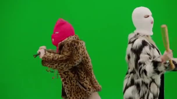两个身穿白色和粉色光环的女人拿着球棒和撬棍跳舞 很滑稽 穿着皮毛外套的女人穿着绿色工作室背景的衣服 时尚潮流的概念 时尚中的女权主义趋势 — 图库视频影像
