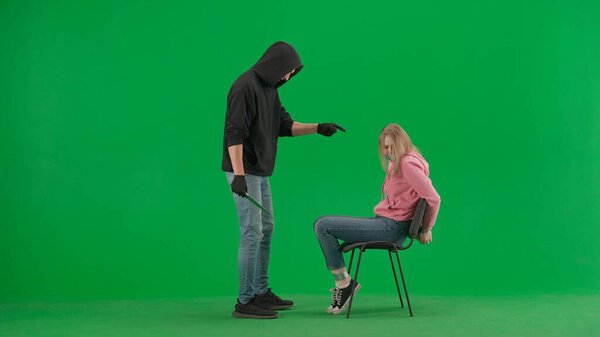 Robbery and criminal concept. Portrait of thief and victim on chroma key green screen background. Girl sitting on chair tied hands and taped mouth man robber with knife threatens her.
