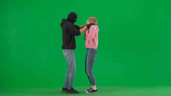 Robbery and crime advertising concept. Portrait of thief and victim on chromakey background of green screen. Male robber in black balaclava mask strangling a girl