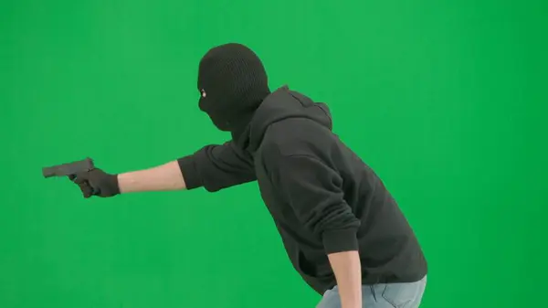 Robbery and criminal concept. Portrait of thief on chroma key green screen background. Man robber in balaclava and hoodie walking holding gun in hand looks around. Side view
