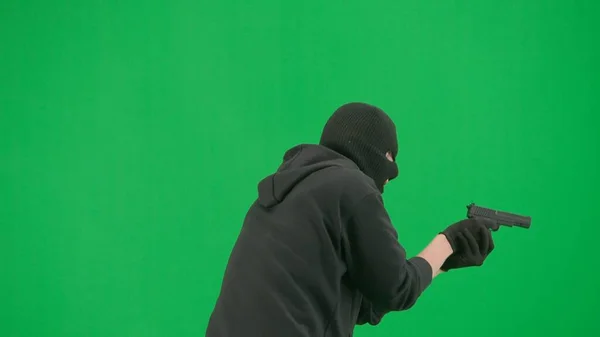 Robbery and criminal concept. Portrait of thief on chroma key green screen background. Man robber in balaclava and hoodie walking holding gun in hand looks around. Back view