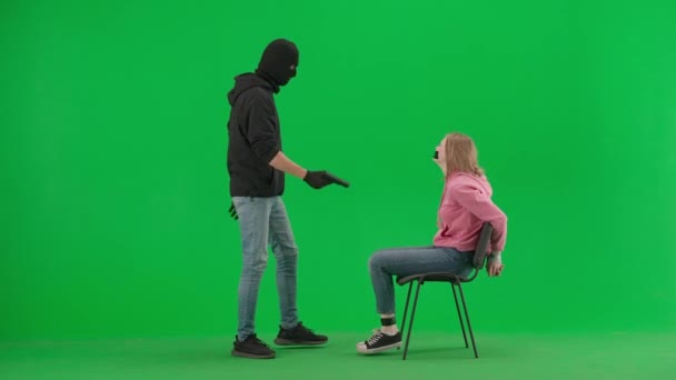 Robbery and criminal concept. Portrait of thief and victim on chroma key green screen background. Girl sitting on chair tied hands and taped mouth man robber with gun threatens her.