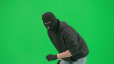 Soygun ve ceza konsepti. Krom anahtar yeşil ekrandaki hırsızın portresi. Kar maskeli ve kapüşonlu bir adam elinde beyzbol sopasıyla yürüyüp etrafa bakıyor. Yarım dönüş.