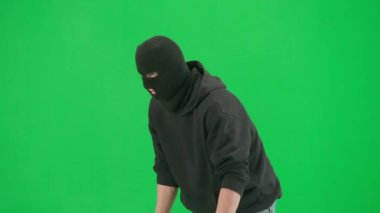 Soygun ve ceza konsepti. Krom anahtar yeşil ekrandaki hırsızın portresi. Kar maskeli ve kapüşonlu bir adam elinde silahla dolaşıyor. Etrafa bakıyor. Yarım dönüş.