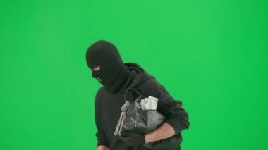 Soygun ve ceza konsepti. Krom anahtar yeşil ekrandaki hırsızın portresi. Kapüşonlu, kot pantolonlu ve kar maskeli bir adam para dolu bir çantayla polisten kaçıyor. Yarım dönüş.