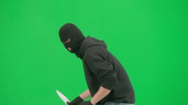 Soygun ve ceza konsepti. Krom anahtar yeşil ekrandaki hırsızın portresi. Kar maskeli ve kapüşonlu bir adam elinde bıçakla dolaşıyor. Etrafa bakıyor. Yan görünüm