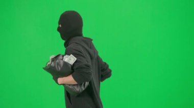 Soygun ve ceza konsepti. Krom anahtar yeşil ekrandaki hırsızın portresi. Kapüşonlu, kot pantolonlu ve kar maskeli bir adam para dolu bir çantayla polisten kaçıyor. Yan görünüm