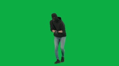 Soygun ve ceza konsepti. Krom anahtar yeşil ekrandaki hırsızın portresi. Kar maskeli ve kapüşonlu bir adam elinde silahla dolaşıyor. Etrafa bakıyor..