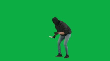 Soygun ve ceza konsepti. Krom anahtar yeşil ekrandaki hırsızın portresi. Kar maskeli ve kapüşonlu bir adam elinde bıçakla dolaşıyor. Etrafa bakıyor. Yarım dönüş.