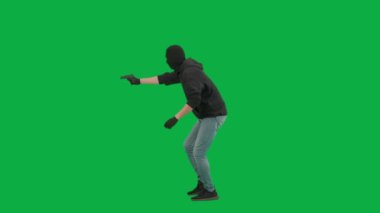 Soygun ve ceza konsepti. Krom anahtar yeşil ekrandaki hırsızın portresi. Kar maskeli ve kapüşonlu bir adam elinde silahla dolaşıyor. Etrafa bakıyor. Yan görünüm