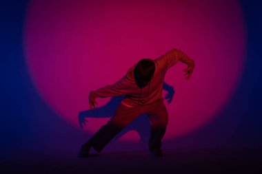 Stüdyoda mavi ışıkla hip hop dansı yapan günlük kıyafetli adam. Dansçı kırmızı ışık çemberinde ve vücut esnekliğini gösteriyor. Tam yükseklik. Modern sokak koreografisi