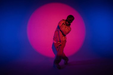 Stüdyoda mavi ışıkla hip hop dansı yapan günlük kıyafetli adam. Dansçı kırmızı ışık çemberinde ve vücut esnekliğini gösteriyor. Tam yükseklik. Modern sokak koreografisi