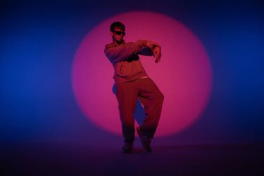 Stüdyoda mavi ışıkla hip hop dansı yapan günlük kıyafetli adam. Dansçı kırmızı ışık çemberinde ve vücut esnekliğini gösteriyor. Tam yükseklik. Modern sokak koreografisi