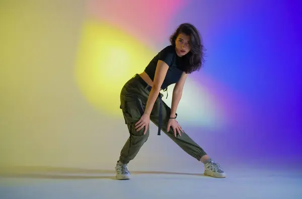Young woman in black clothes posing in studio with gradient blue, yellow, pink neon light. Dancer demonstrates elements of jazz funk style dance. Contemporary choreography
