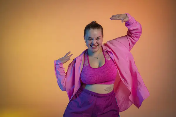 Young woman in a pink shirt dances to jazz funk rhythms against the backdrop of a orange studio. The girl confidently expresses her personality through dance, genuinely enjoying the music
